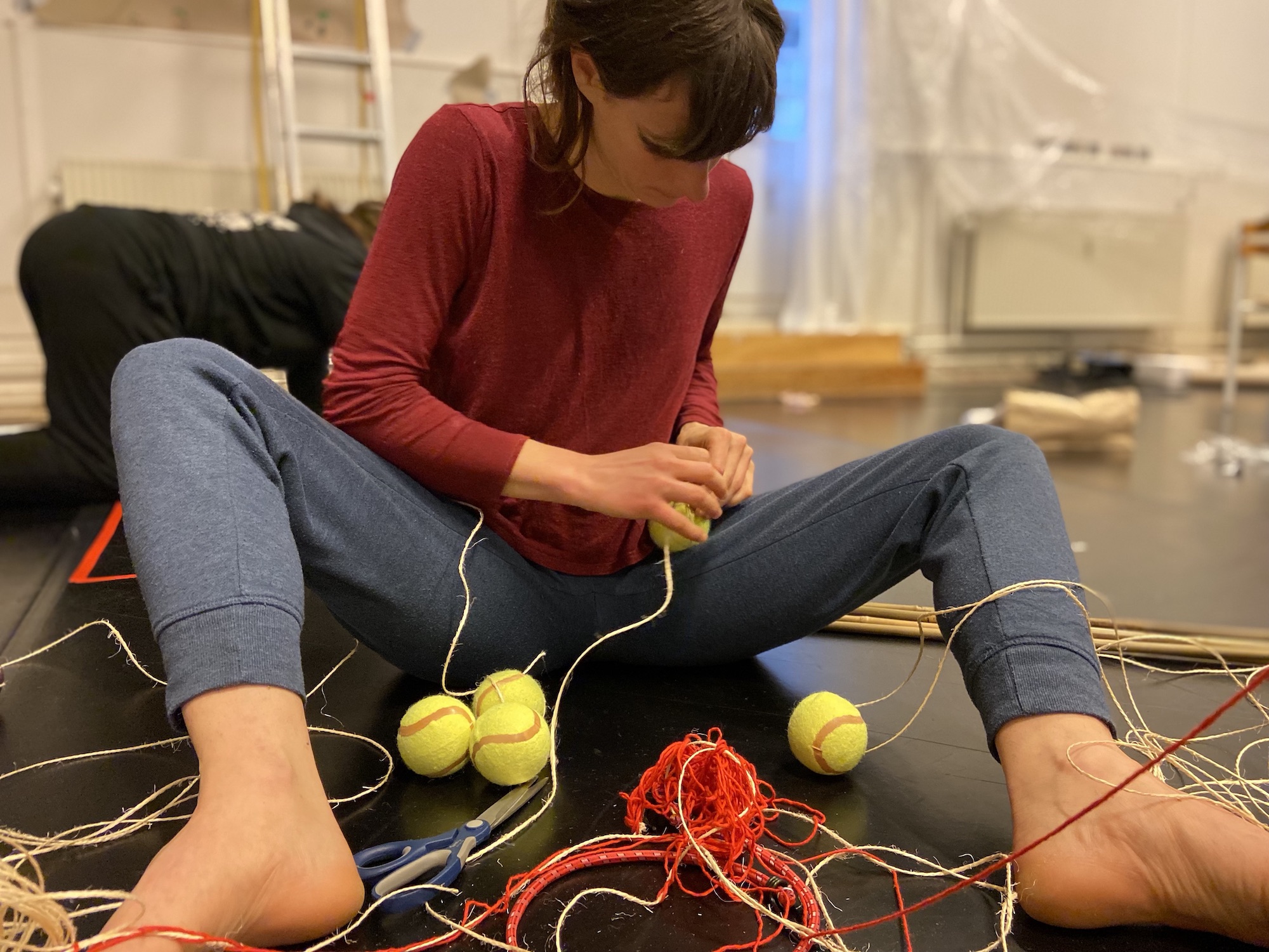 A participant sits on the floor with their knees spread out, making something involving tennis balls and thick string