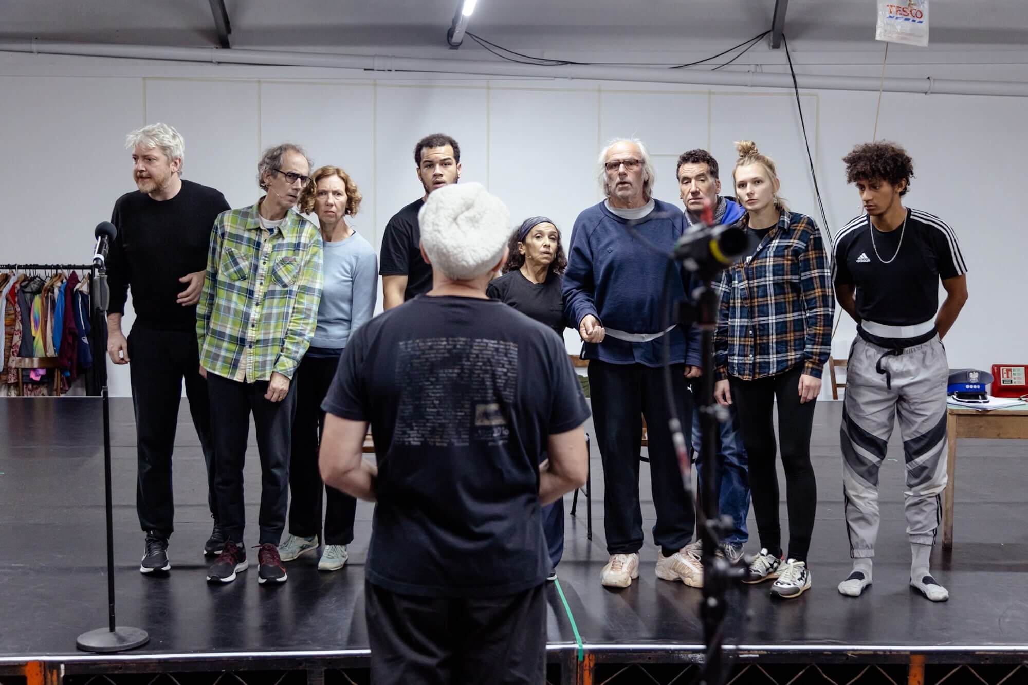 In rehearsals, the cast stand closely in a line staring suspiciously at the camera. Simon McBurney's back is to the camera.