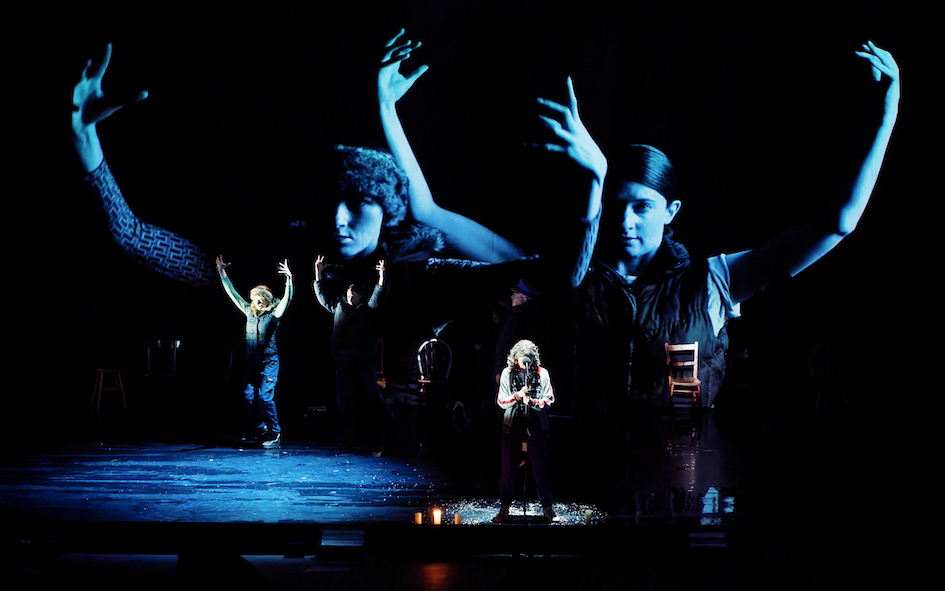 An older woman stands at a microphone in the centre, two actors representing deer stand to her left. The two actors are projected so that they are large across the back of the stage, standing and waiting.