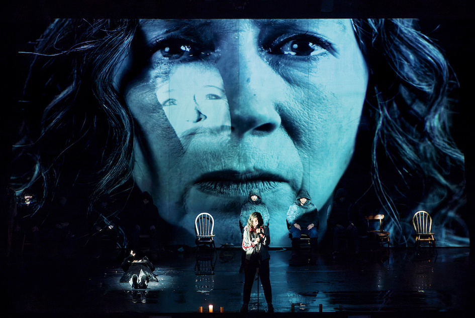 A woman's face is projected in cool blue tones across the back of the stage and the real woman is seen, tiny, in the foreground.