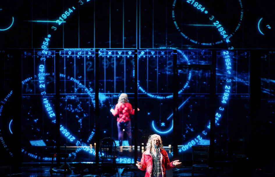 Astrological symbols are beamed in large blue circles and charts across the back of the stage, as an older woman speaks direct to the audience to the audience wearing a red coat.