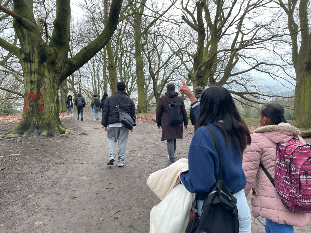 The backs of a group of students walking through a wood, while the trees are bare. One student points in the foreground.