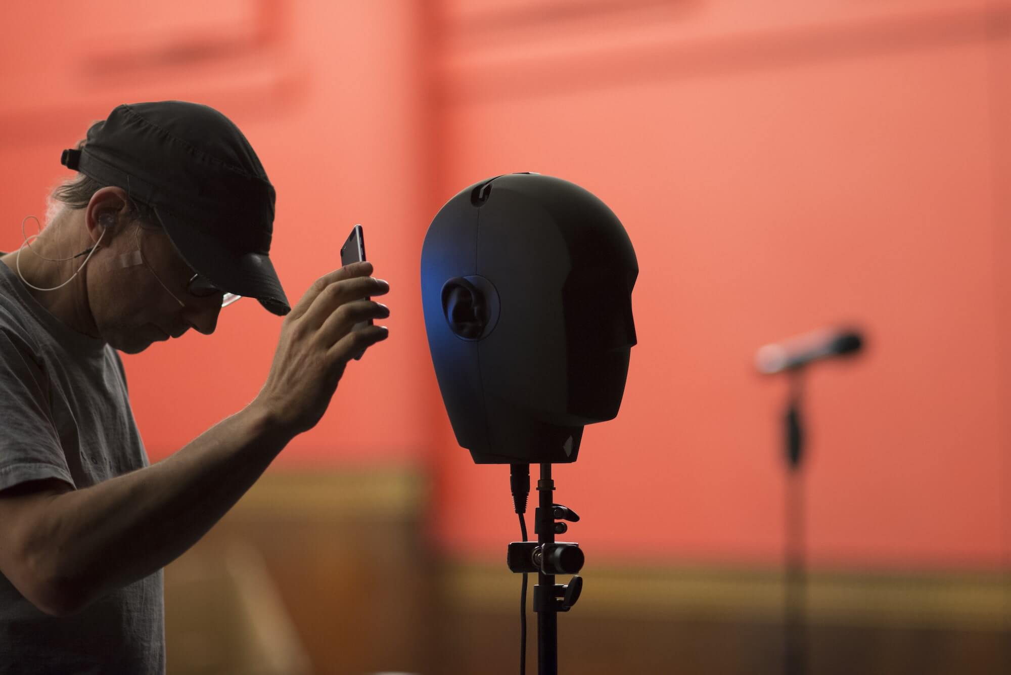 In rehearsals for The Encounter, Simon McBurney bows his head and holds up his phone to the iconic bianural head in a coral-shaded room
