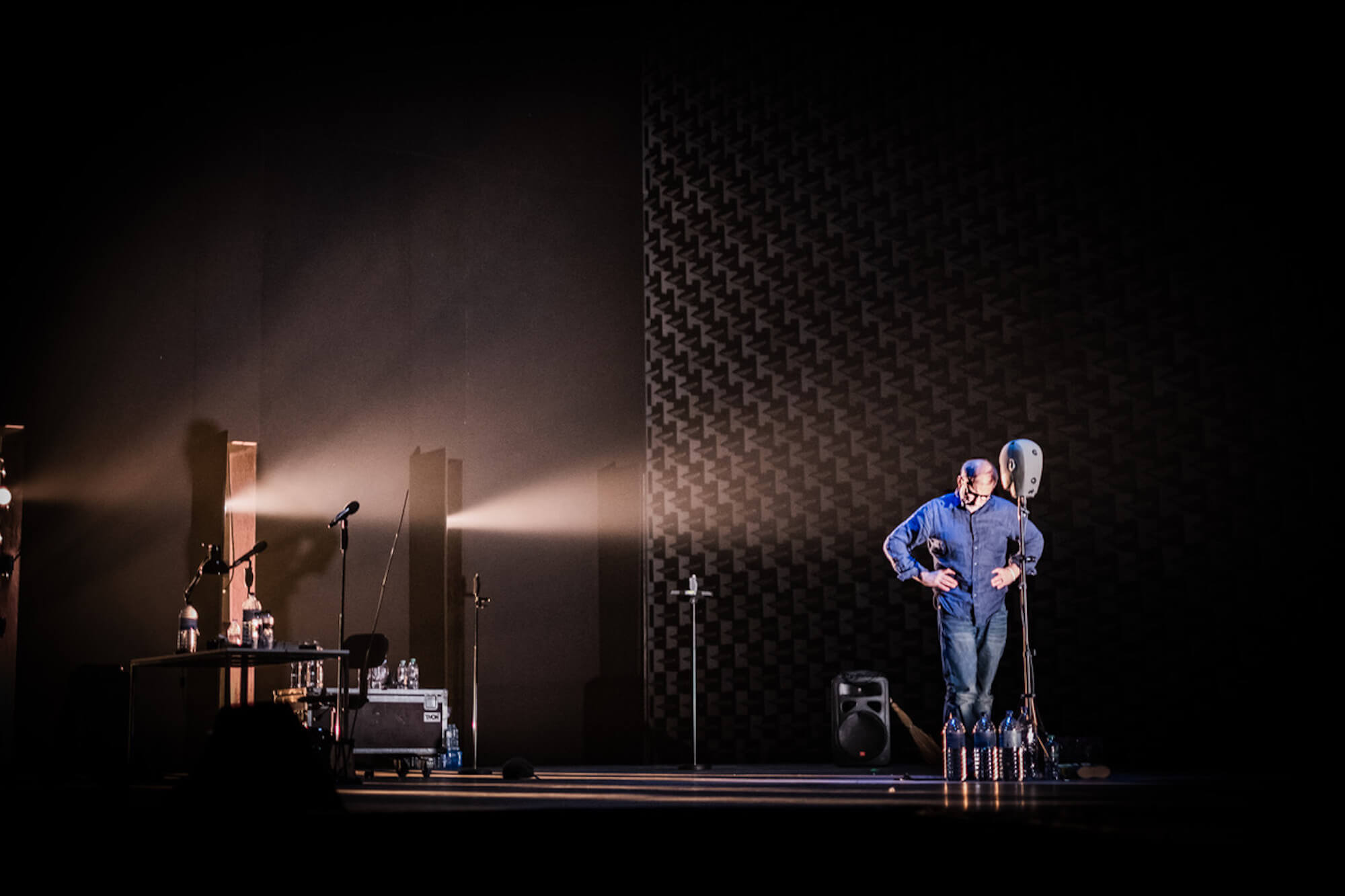 In a dark, black stage, with two single streams of light shining from the back, Simon McBurney stands looking down with his hands on his hips, while the stage is arrayed with many water bottles.