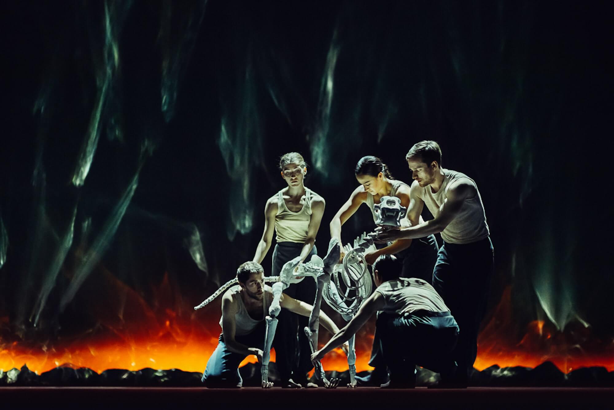 A group of serious looking dancers manipulate a skeleton with four legs and a long tail, with a backdrop of destruction and fire behind them.