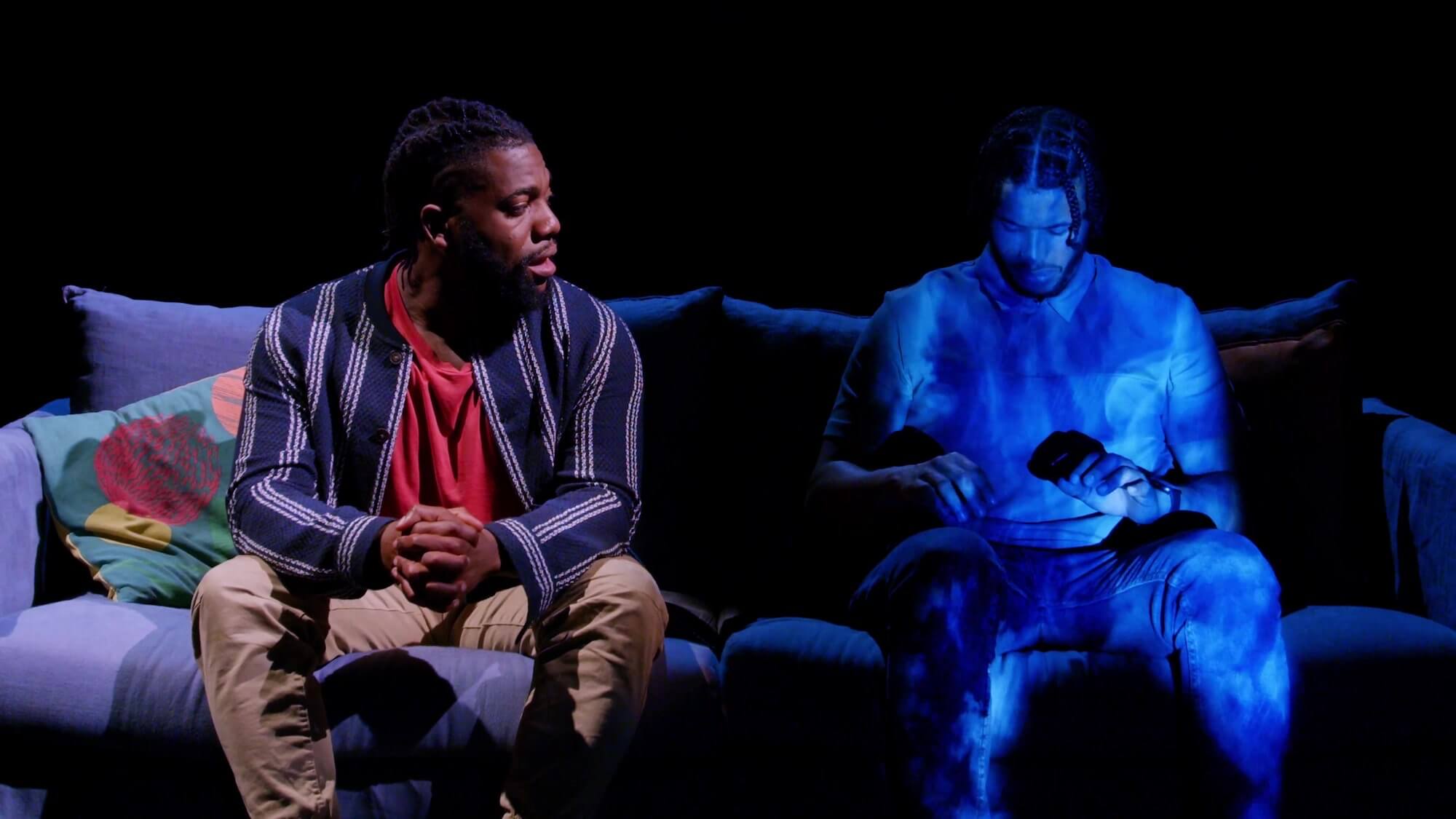 Fehinti Balogun sits on a sofa, talking seriously to a hologram person sitting next to him