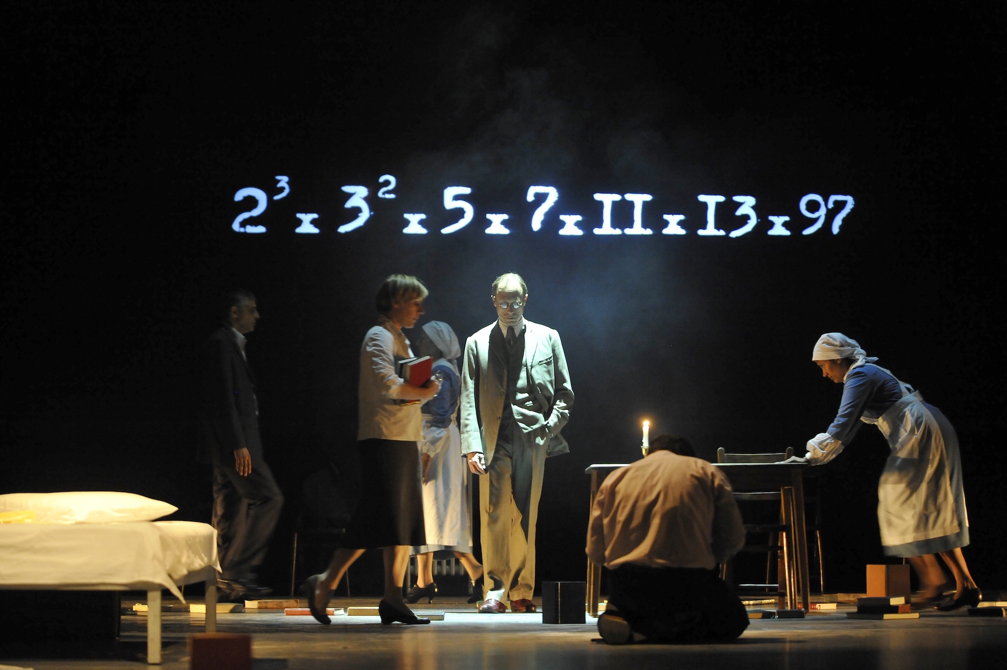 From A Different Number, multiple actors move across the stage, some dressed in office attire, some dressed as nurses, while a mathematical equation is projected on the black back wall.