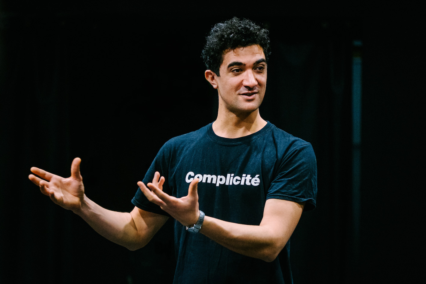 A facilitator speaks calmly, gesturing both arms to the left of the picture, while wearing a Complicté branded tea shirt