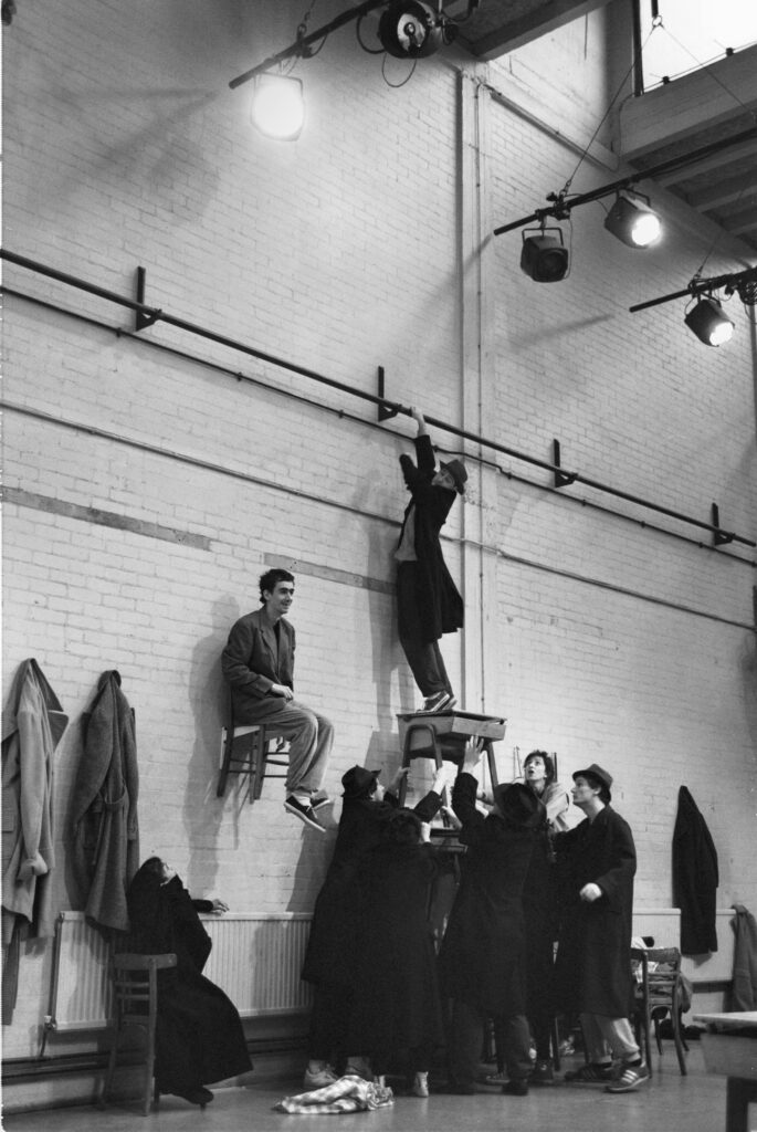 The company gather around an actor, who is standing on several stacked desks reaching away from them, while another actor sits on a chair attached half way up the wall.