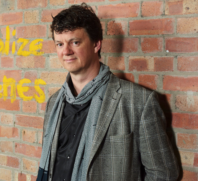 Tom is pictured standing straight against a brick wall with some yellow writing on it, looking at the camera with a gentle air.