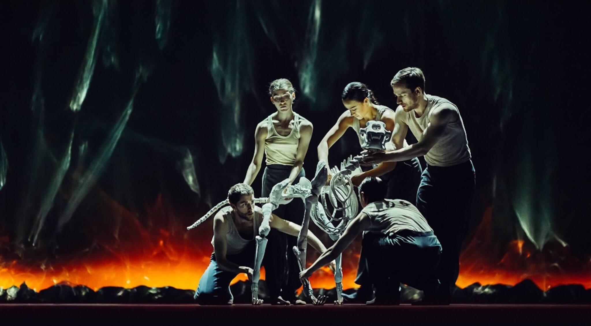 In Figures in Extinction, a group of serious looking dancers manipulate a skeleton with four legs and a long tail, with a backdrop of destruction and fire behind them.