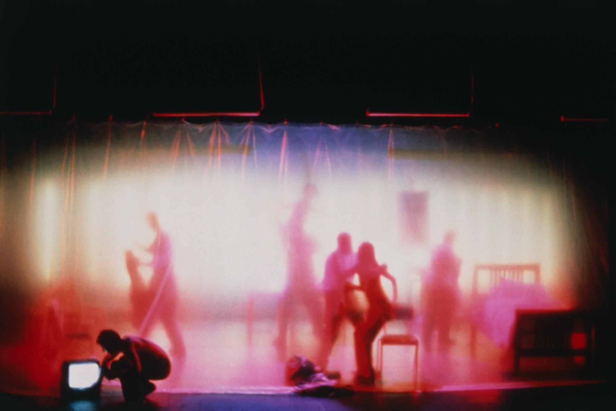 Shadow-like actors bathed in a rich pink move around the stage amongst chairs and a bed with a sheer curtain in front of them.