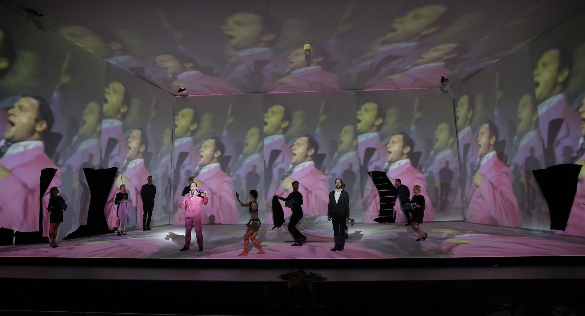 Multiple members of the cast look at an actor singing in a pink suit, who is projected on the back of the stage multiple times, closing in on their face