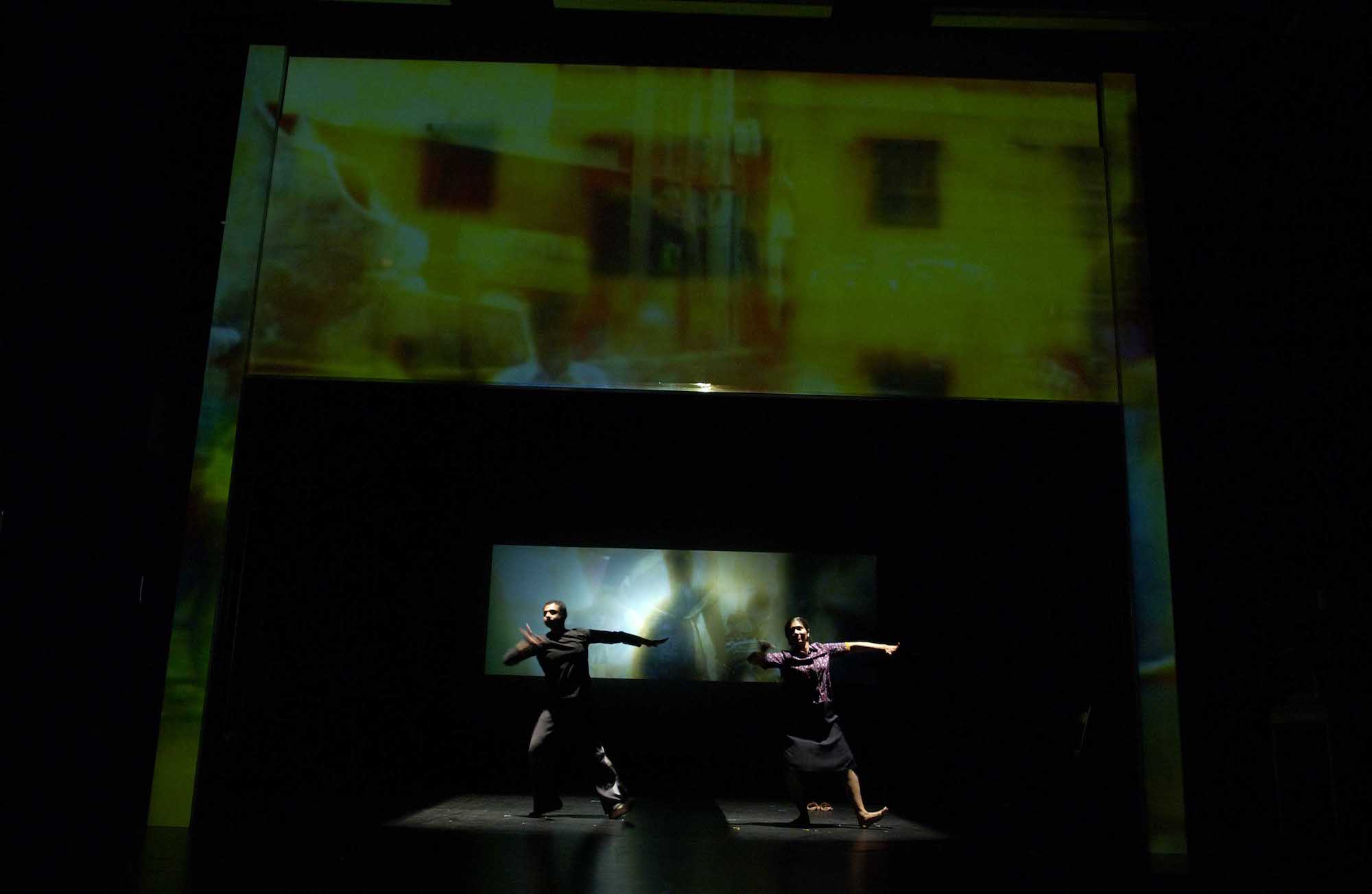 Two actors move in unison on a dark stage, with their left feet raised with the heel on the ground and their left arms out wide, while their right arm is tucked