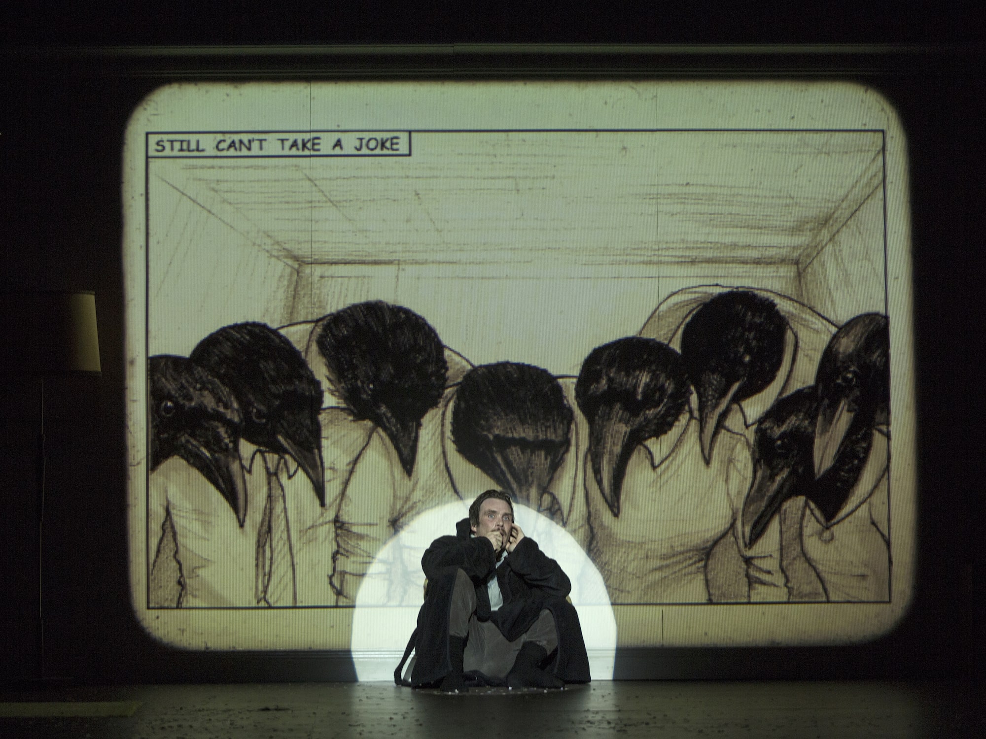 A frightened man sits in a spotlight wearing a dressing gown, while an image of multiple crows with human bodies look down on him. The text reads 