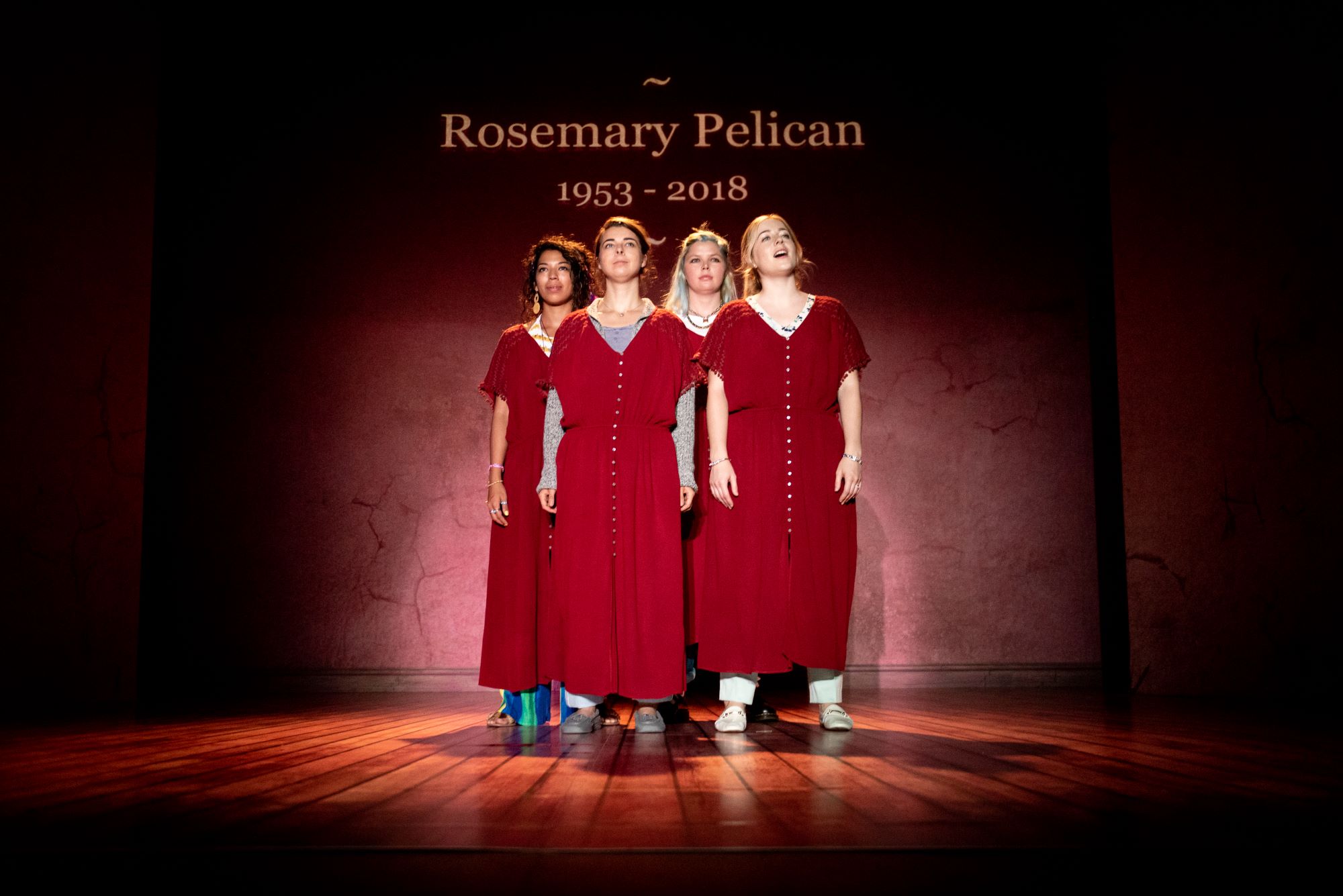 The four sisters stand in formation in red dresses, memorialising, while the text beamed behind them reads 