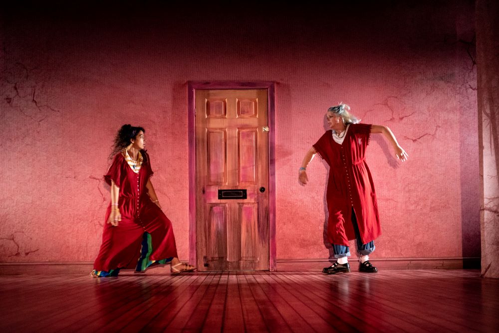 Two sisters wearing red dresses stand agitatedly either side of a door