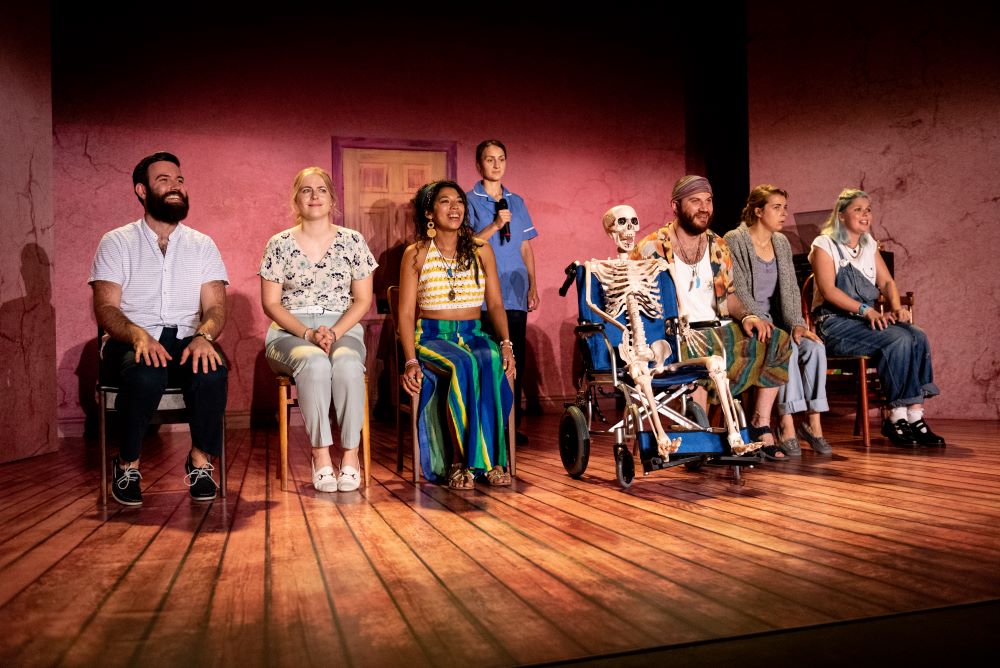 The full cast stand on seats grinning manically, while in the middle of them is a skeleton in a wheelchair with a nurse behind them
