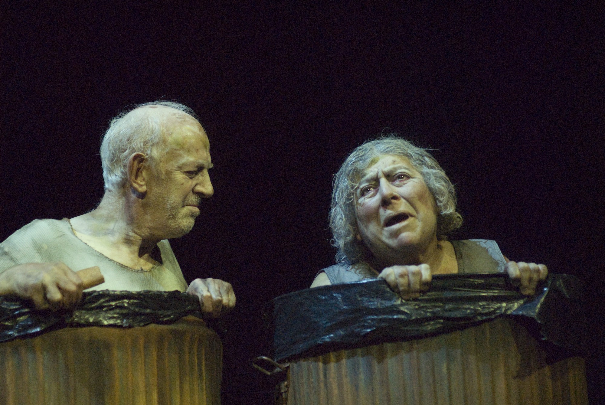 Two actors stand in metal bins with bin bags in them, lamenting their lives