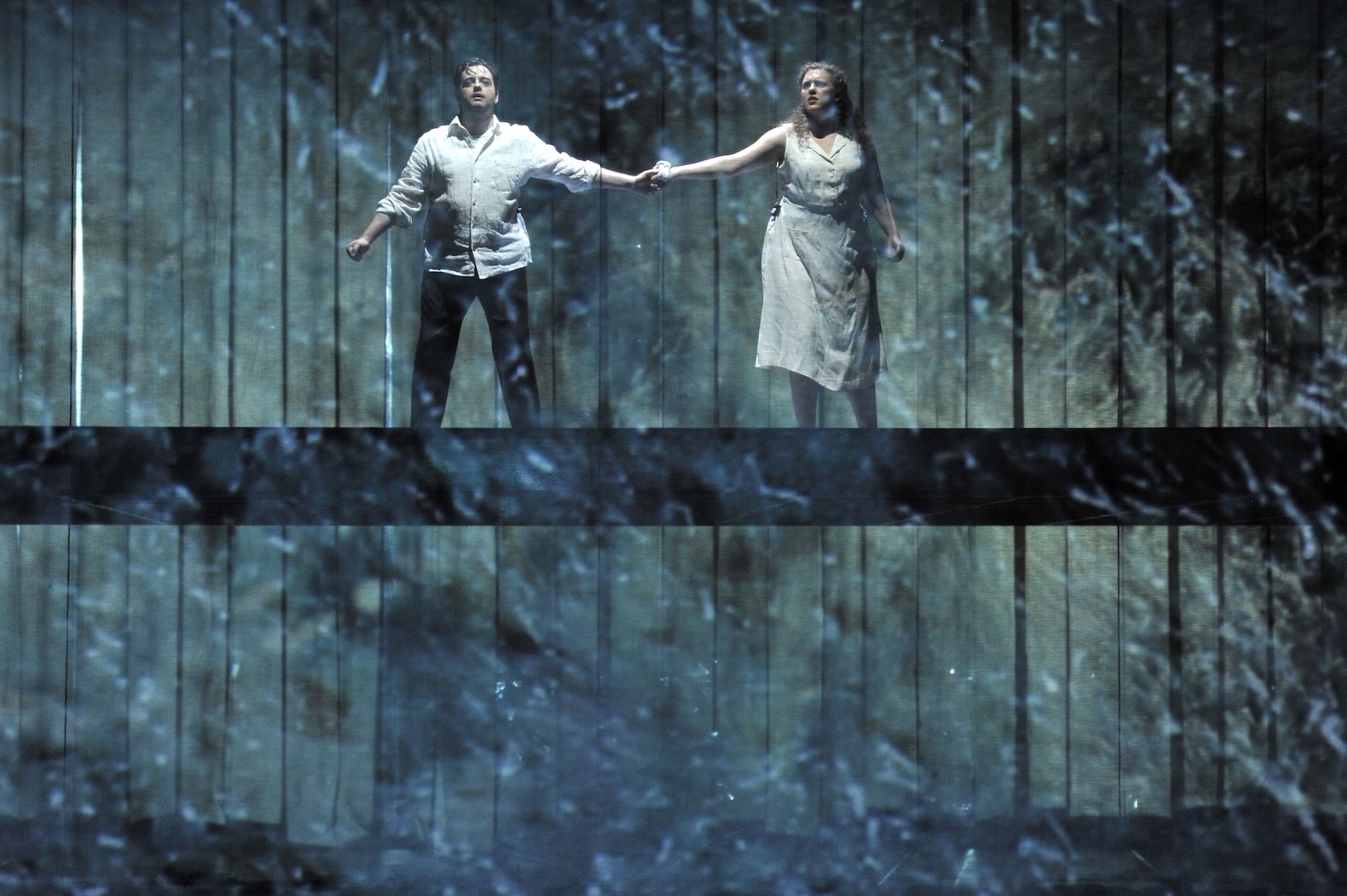 A pair of lovers hold each other's hands as they look at a threat, while standing on a raised bridge across the stage