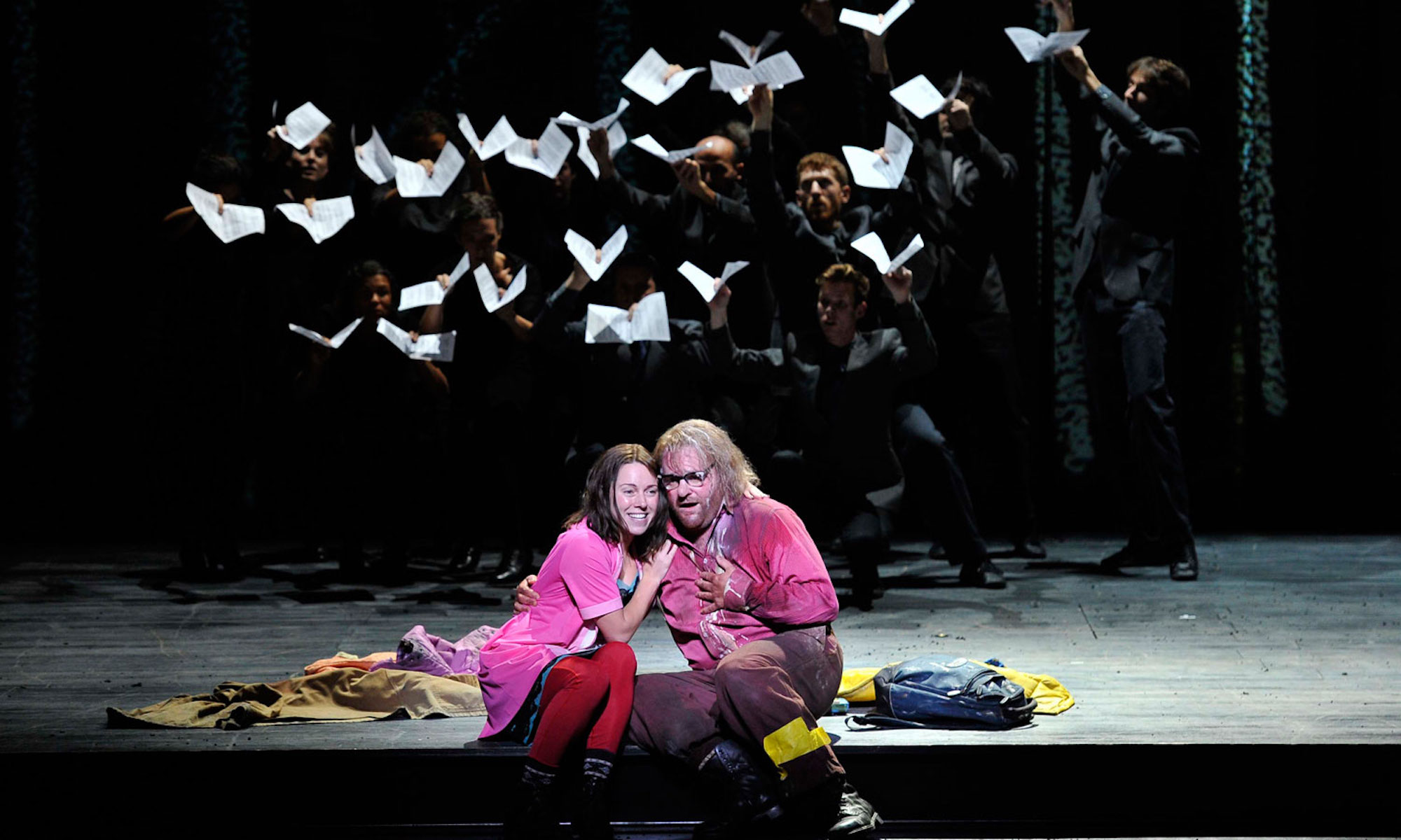 Two actors in pink cuddle together romantically, while behind them, the other actors use books to mimic a bird flying