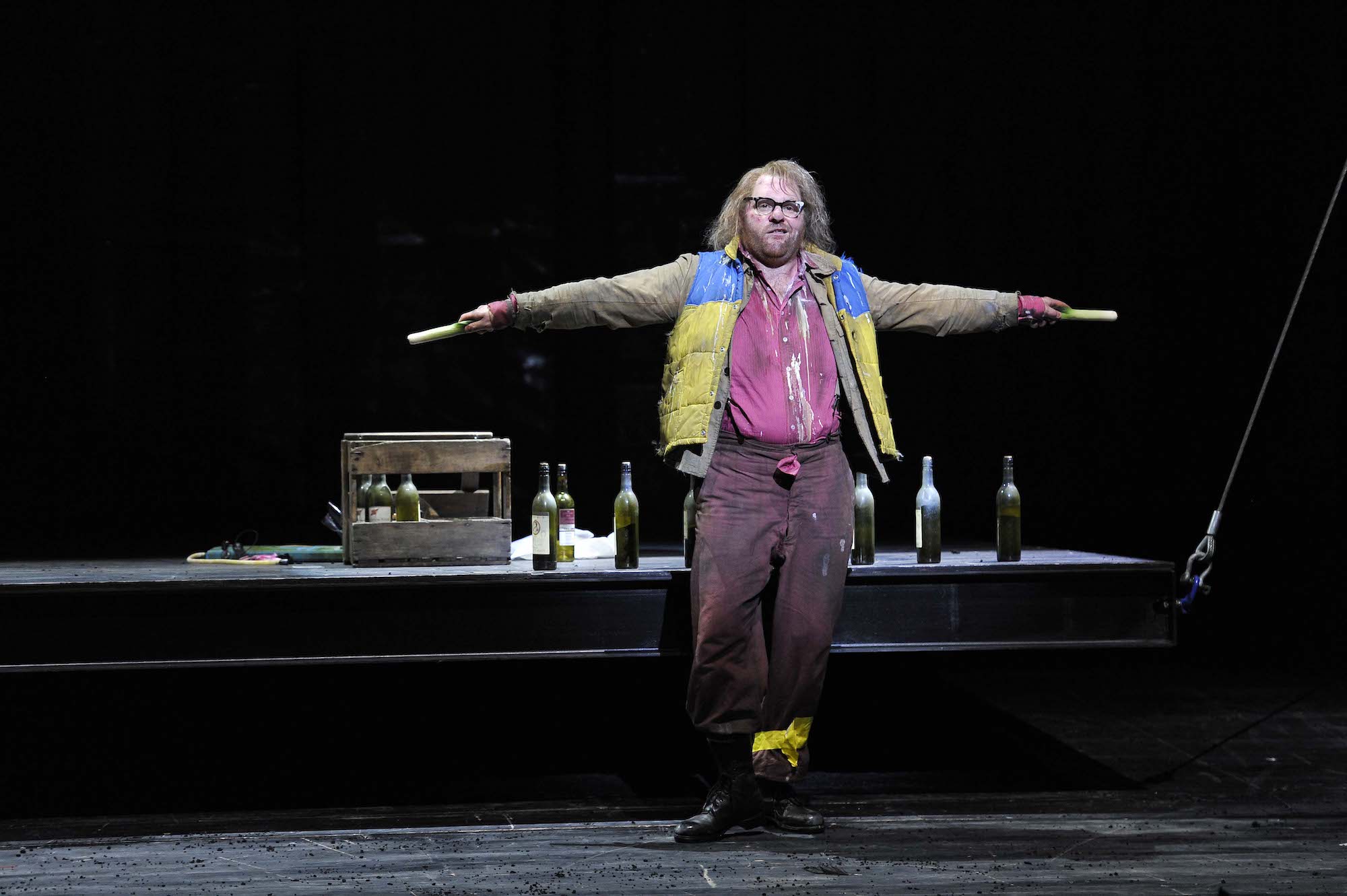 An actor in patched up and pain-splattered clothing makes a comic speech, while being surrounded by empty wine bottles