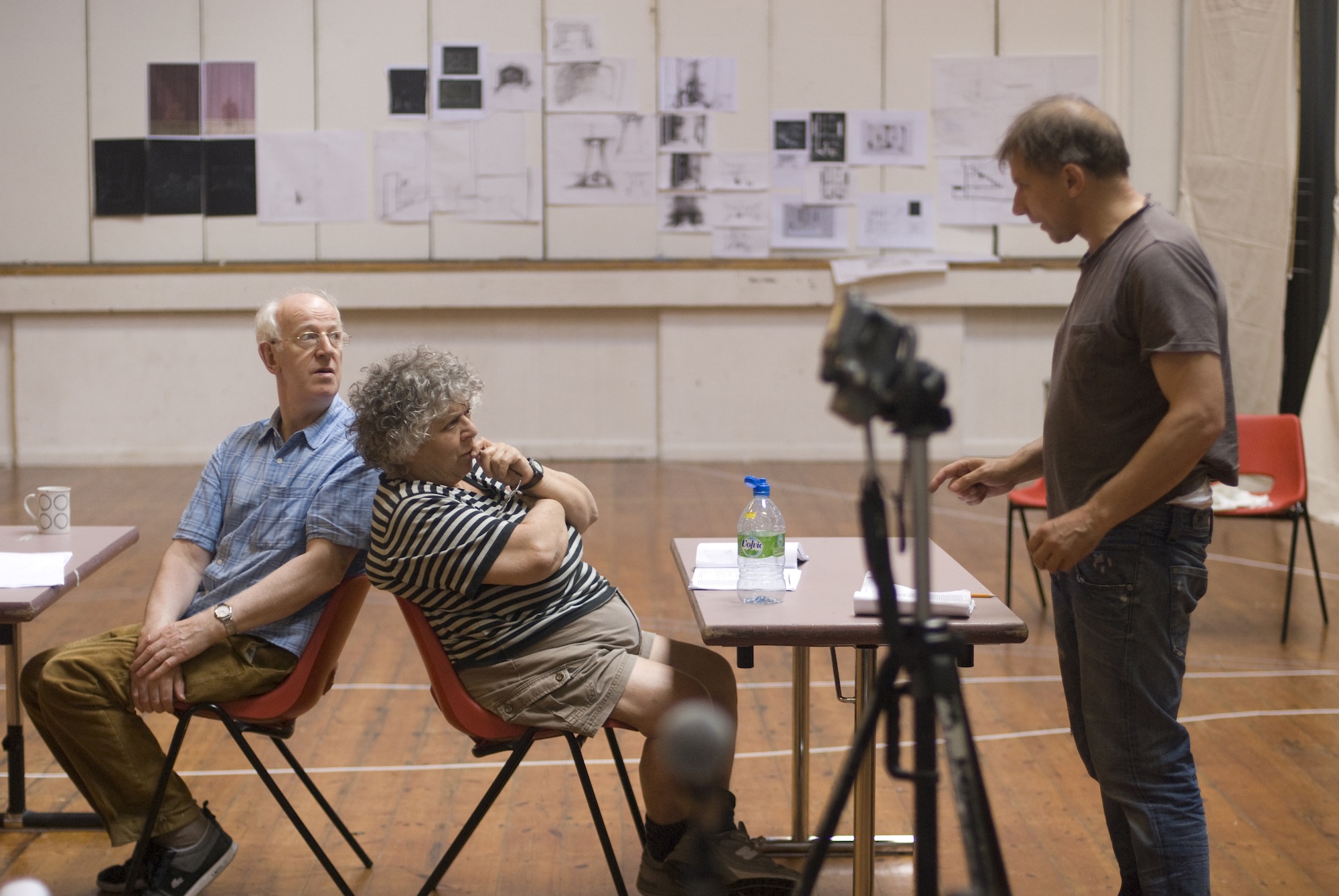 In rehearsals, two sitting actors talk to a standing director, all looking thoughtful