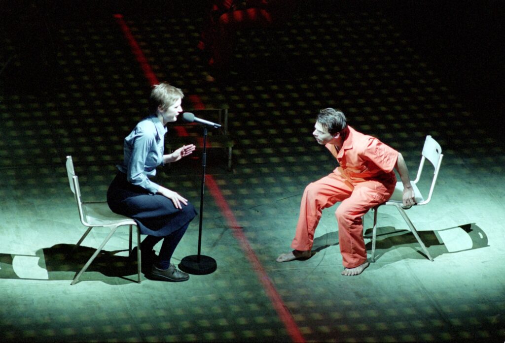 The actor on the left leans forward on their chair, speaking desperately, while the actor on the right in an orange prison uniform, faces them and leans off their chair frantically.