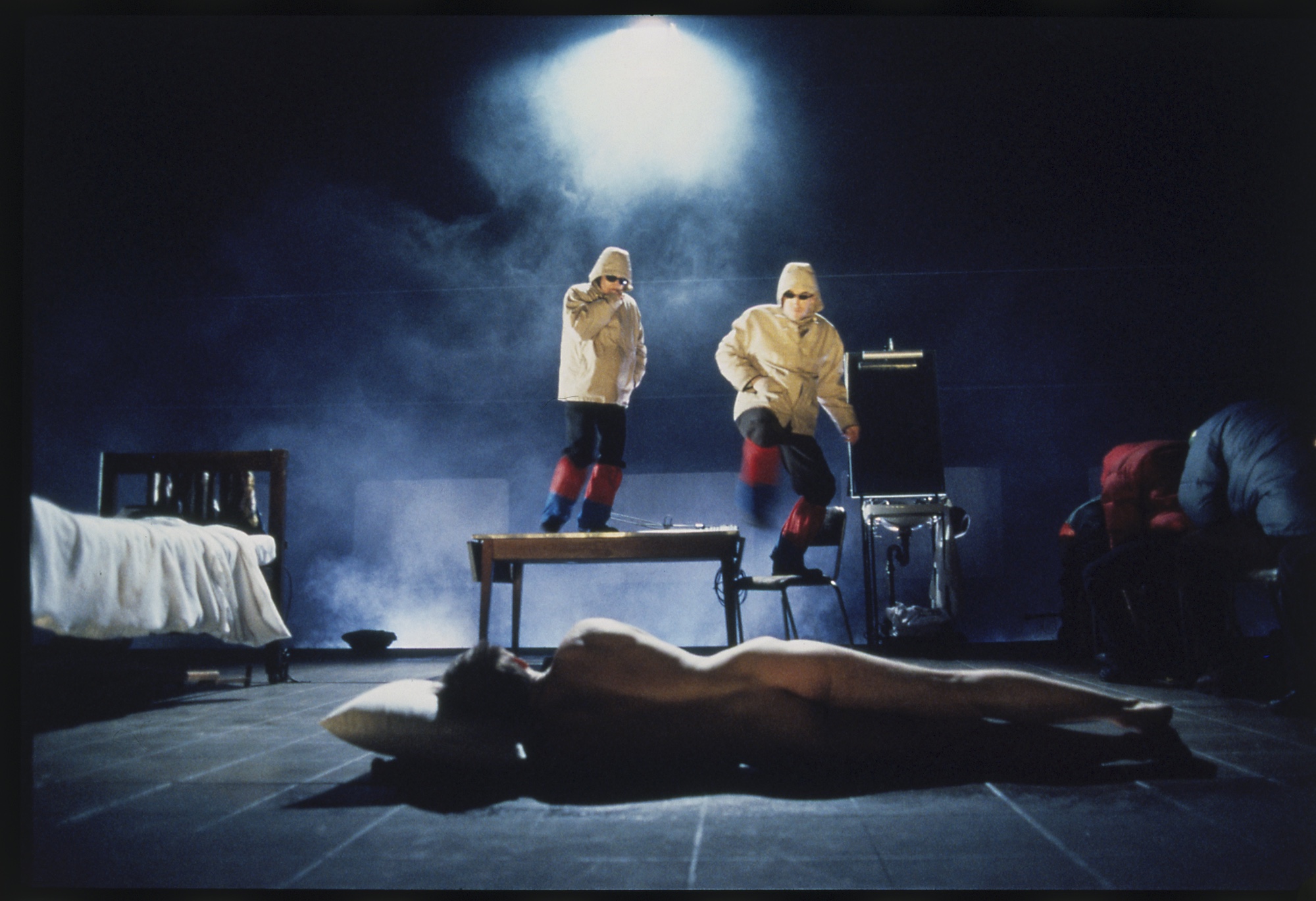 Two actors in yellow raincoats stand on a table and a chair staring intensely a naked actor lying on the floor, who has their back to us.