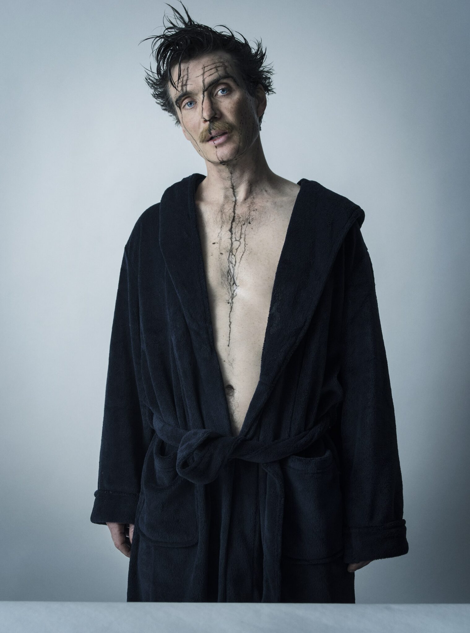 An actor wearing a dressing gown with the front open, showing their bare chest. They have ink over their face, running down their body, and dishevelled hair.