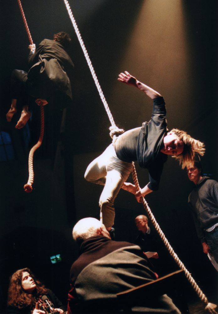 Two actors climb and tumble down ropes hanging from the ceiling, while several other actors watch them