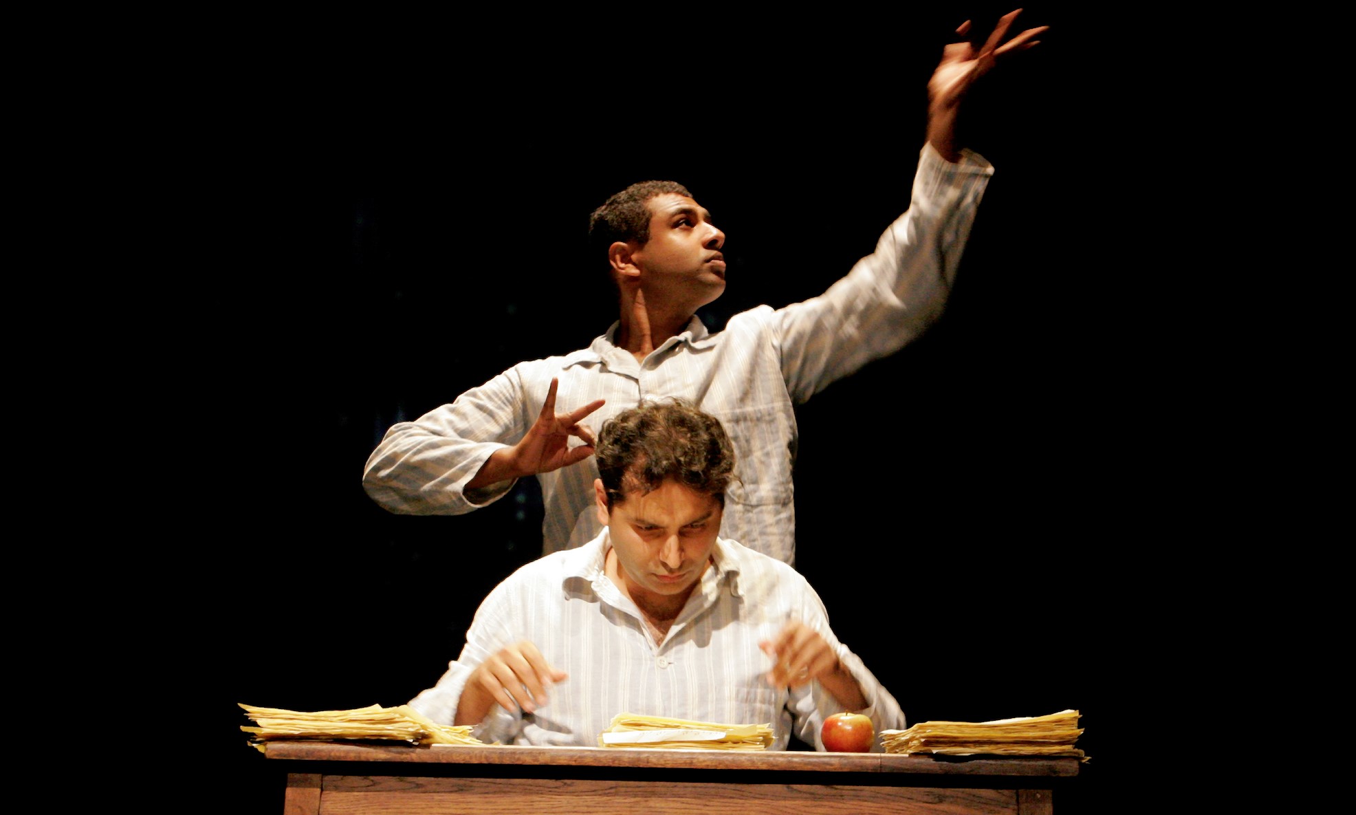 In A Disappearing Number, an actor stares seriously at the pile of papers on the desk, while another actor behind reaches a hand ponderously upwards.