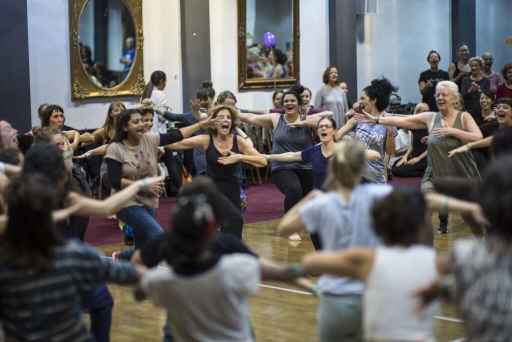 A large group of workshop participants move in a circle joyfully, placing one arm outwards and the other across their chests.