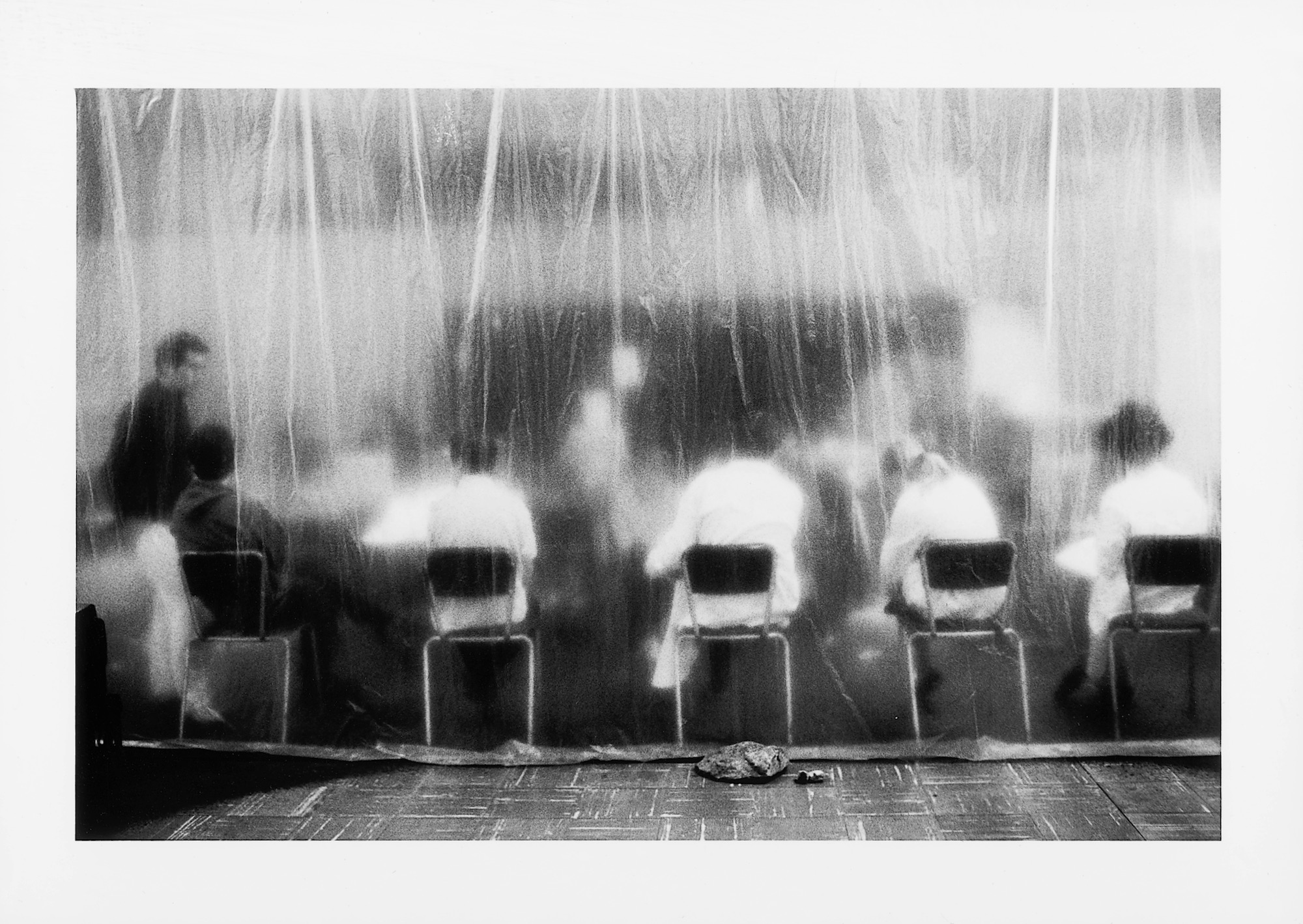 In a black and white photo, a group of shadow-like actors, mostly wearing white coats and sitting on dark chairs, are seen through a sheer plastic curtain.