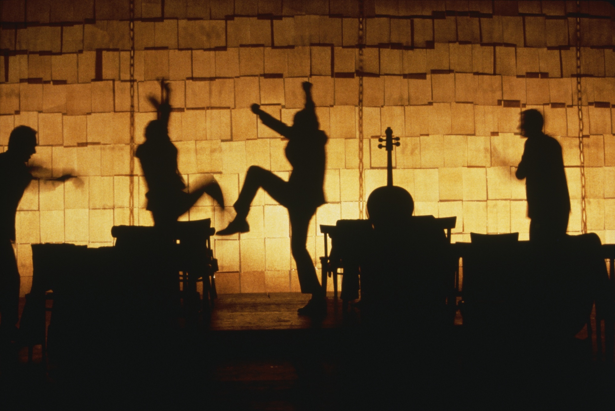 Four shadow-like actors dance in front of a glowing yellow and orange backdrop, amongst shadow-like desks, chairs and a cello.